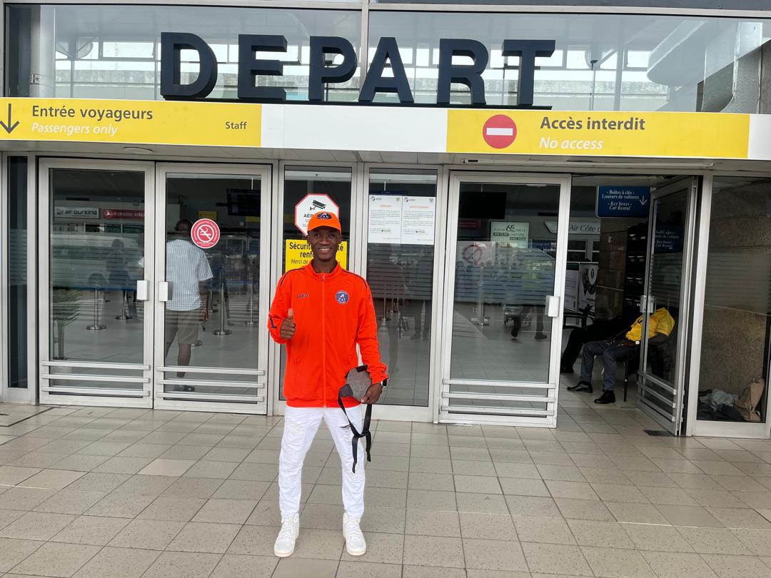 Joueur de JR MIROOL CI souriant devant l'entrée des voyageurs à l'aéroport